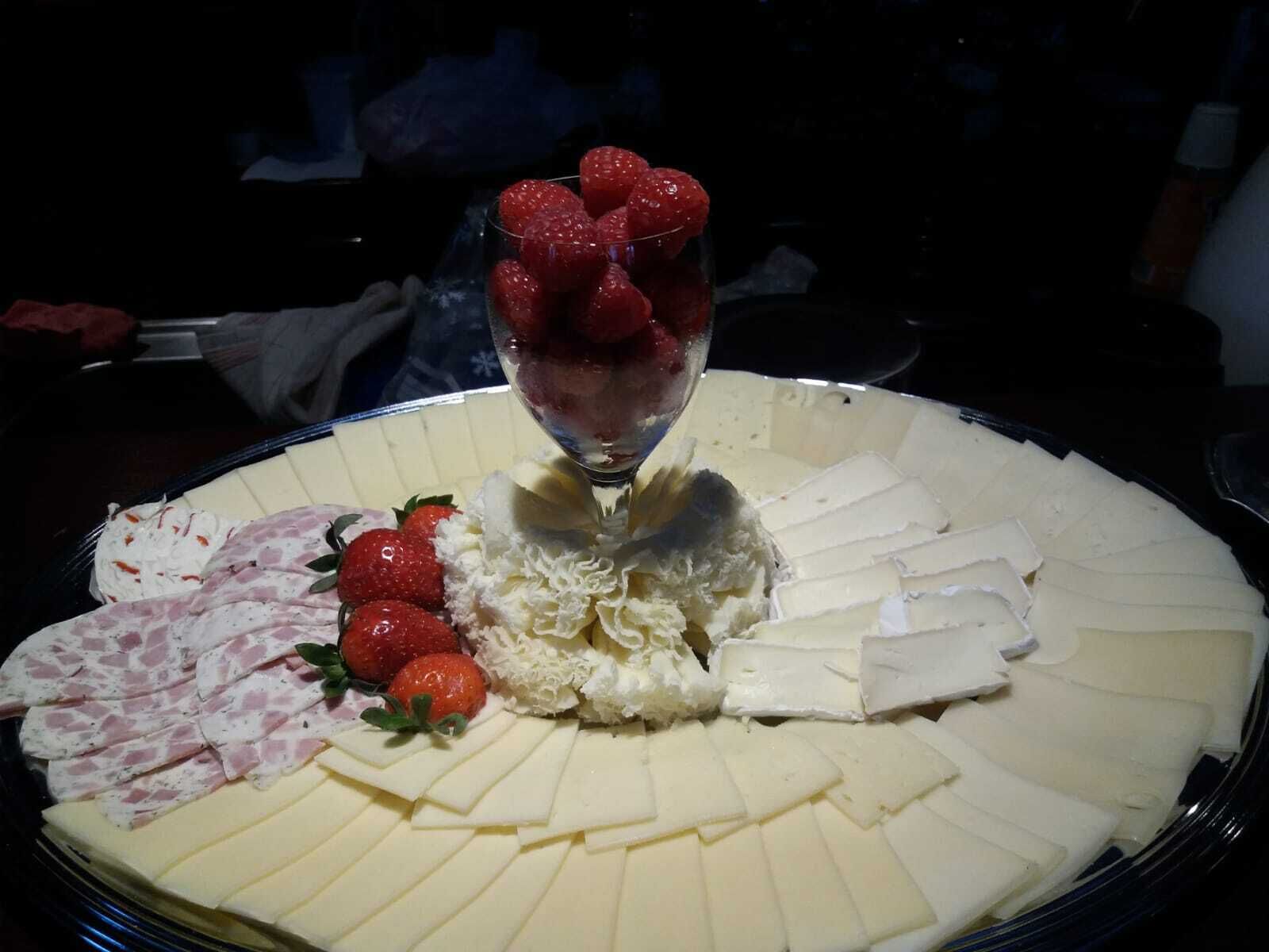 Un plateau garni d’un assortiment de fromages, notamment une portion de brie, des tranches de camembert et une sélection de charcuteries, accompagné de fraises fraîches et d’un verre de vin rouge.