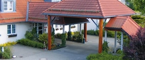 A covered patio area with a tiled roof, surrounded by greenery and a paved walkway leading to a building with a red-tiled roof. - Business Hotel Siebeneichen GmbH (Meissen)