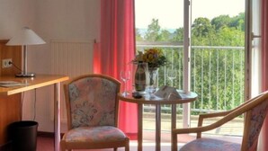 A hotel room with a wooden desk, a television, a chair, a small round table with a vase of flowers, and a balcony with a view of greenery. - Business Hotel Siebeneichen GmbH (Meissen)