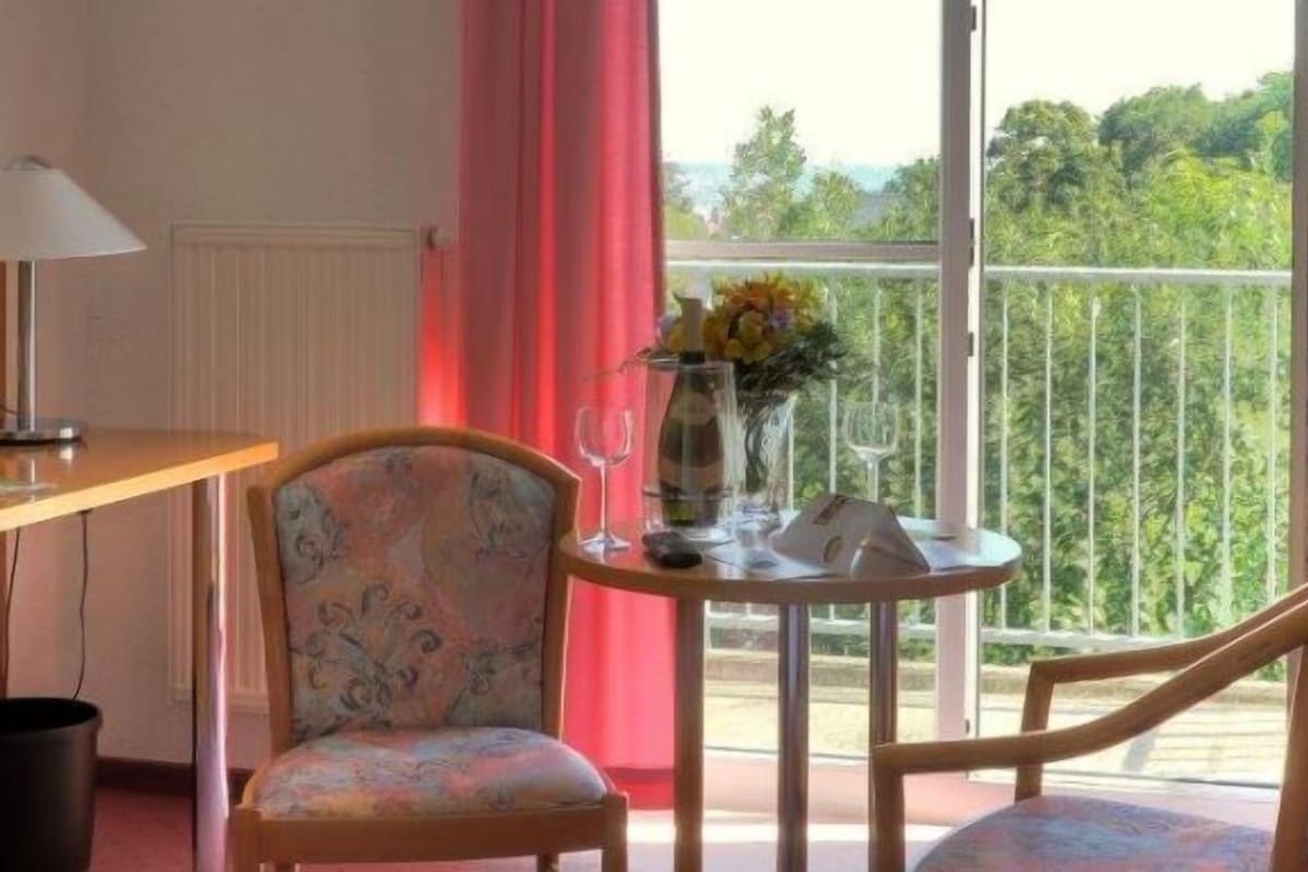 A hotel room with a wooden desk, a television, a chair, a small round table with a vase of flowers, and a balcony with a view of greenery.