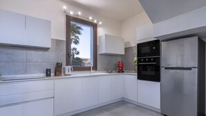 Eine moderne Küche mit weißen Schränken, einem Edelstahlkühlschrank, einem eingebauten Backofen und einem Fenster mit Blick auf Palmen.