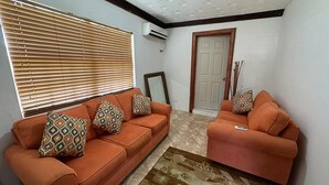 Una sala de estar con dos sofás naranja, cojines estampados, una puerta de madera y piso de baldosas.