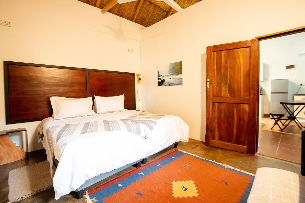 A bedroom with a wooden headboard, a bed with white bedding, a colorful rug, and a wooden door.