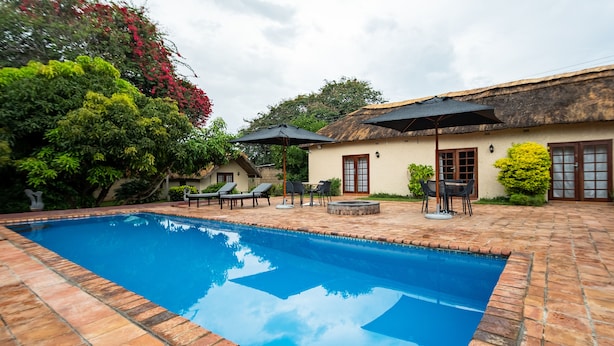 Piscina com espreguiçadeiras e guarda-sóis, rodeada por um pátio de tijolos e um edifício com telhado de palha.