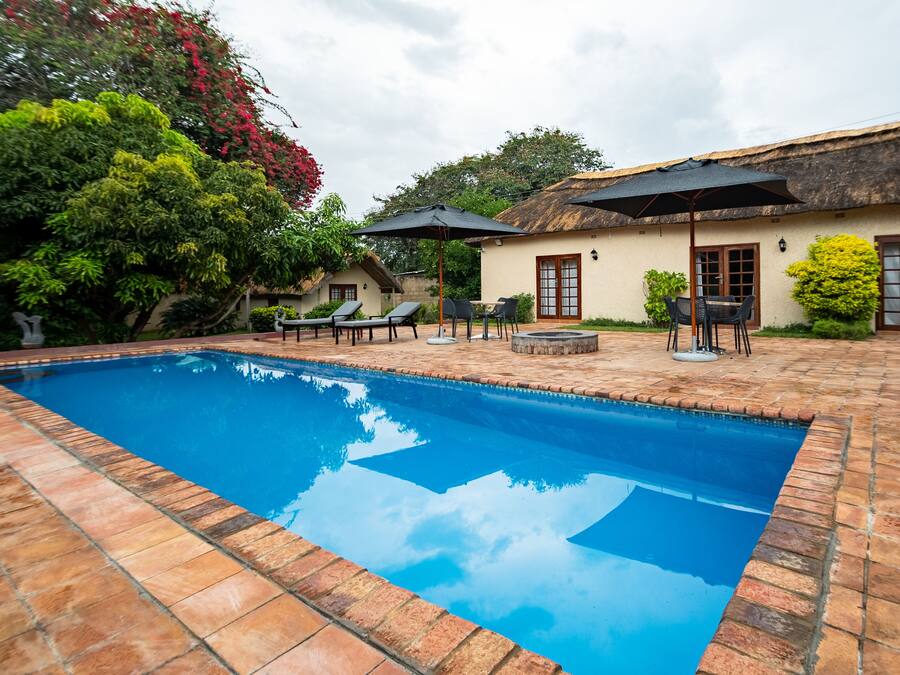 Una piscina con sillas de descanso y sombrillas, rodeada por un patio de ladrillo y un edificio con techo de paja.