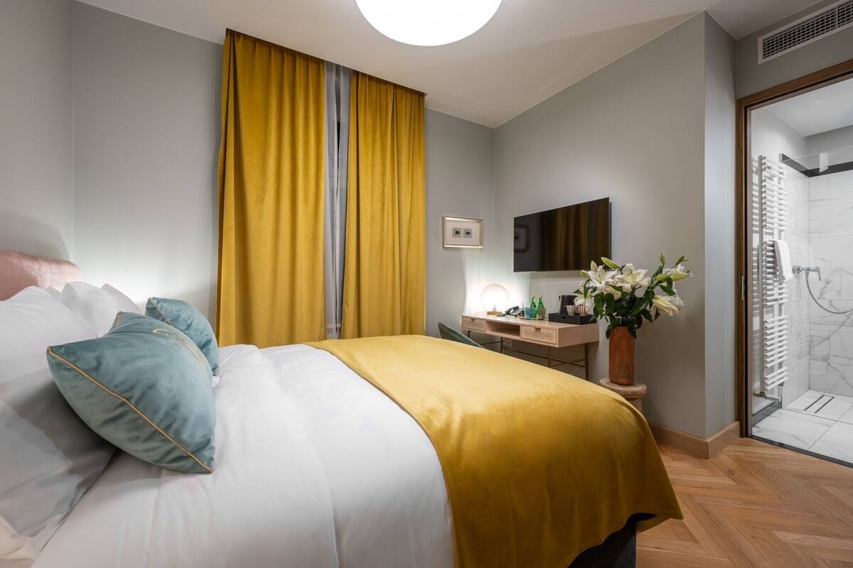 A hotel room with a bed, a desk with a lamp, a vase with flowers, and a shower area.