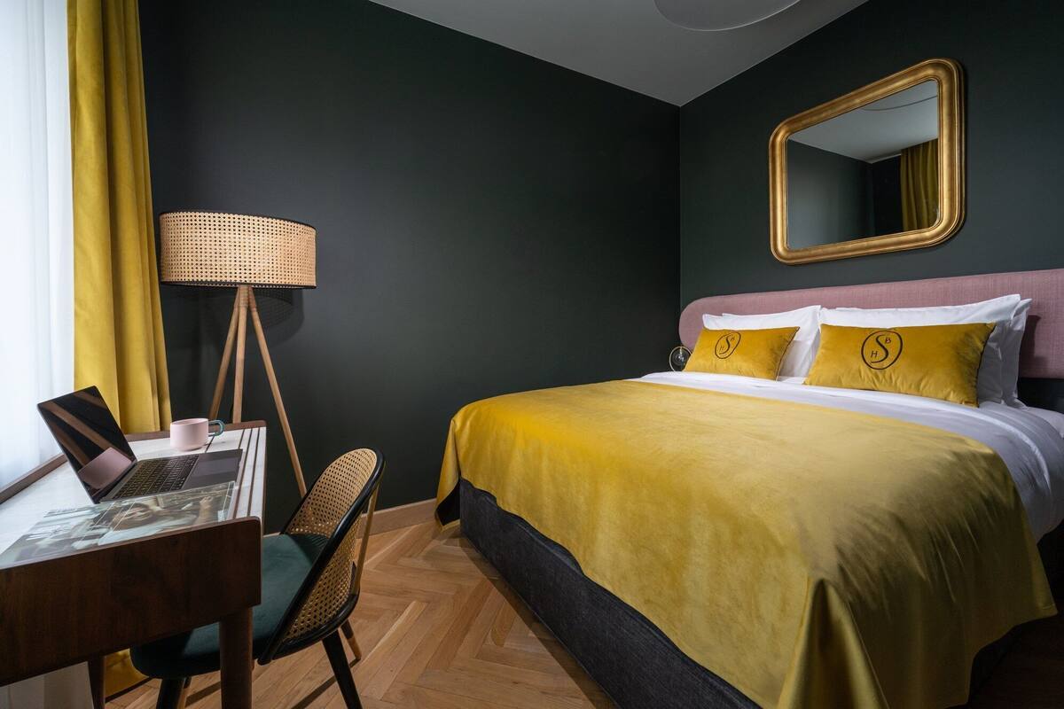 A hotel room with a bed, a desk with a laptop, a chair, a lamp, and a mirror.