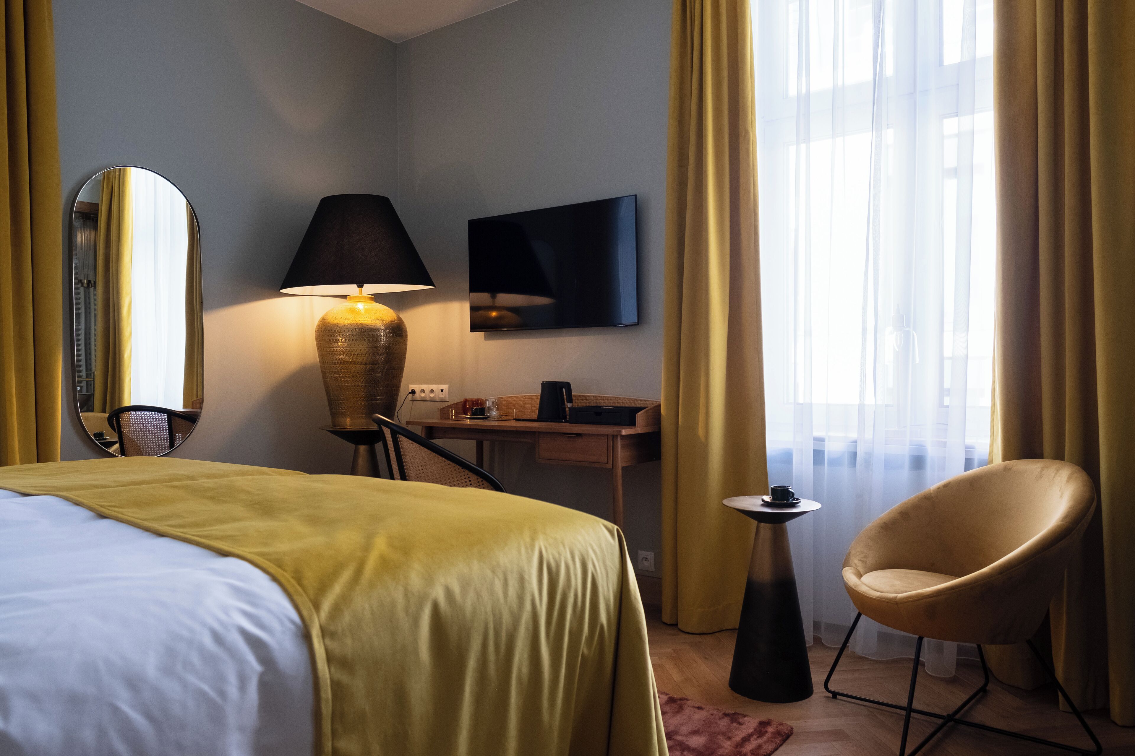 A hotel room with a bed, a desk, a chair, a lamp, a TV, and a window with curtains.