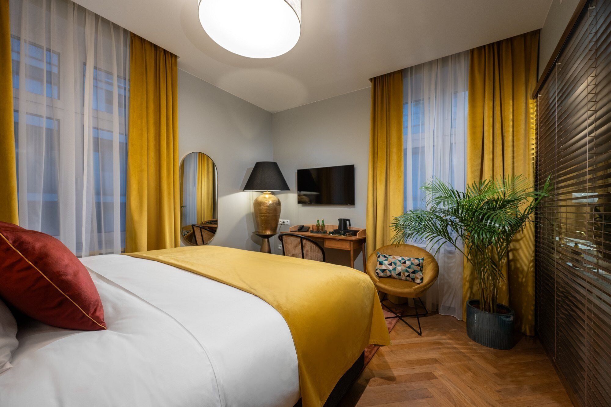 Quarto de hotel com cama, escrivaninha, cadeira, televisão e planta em vaso.