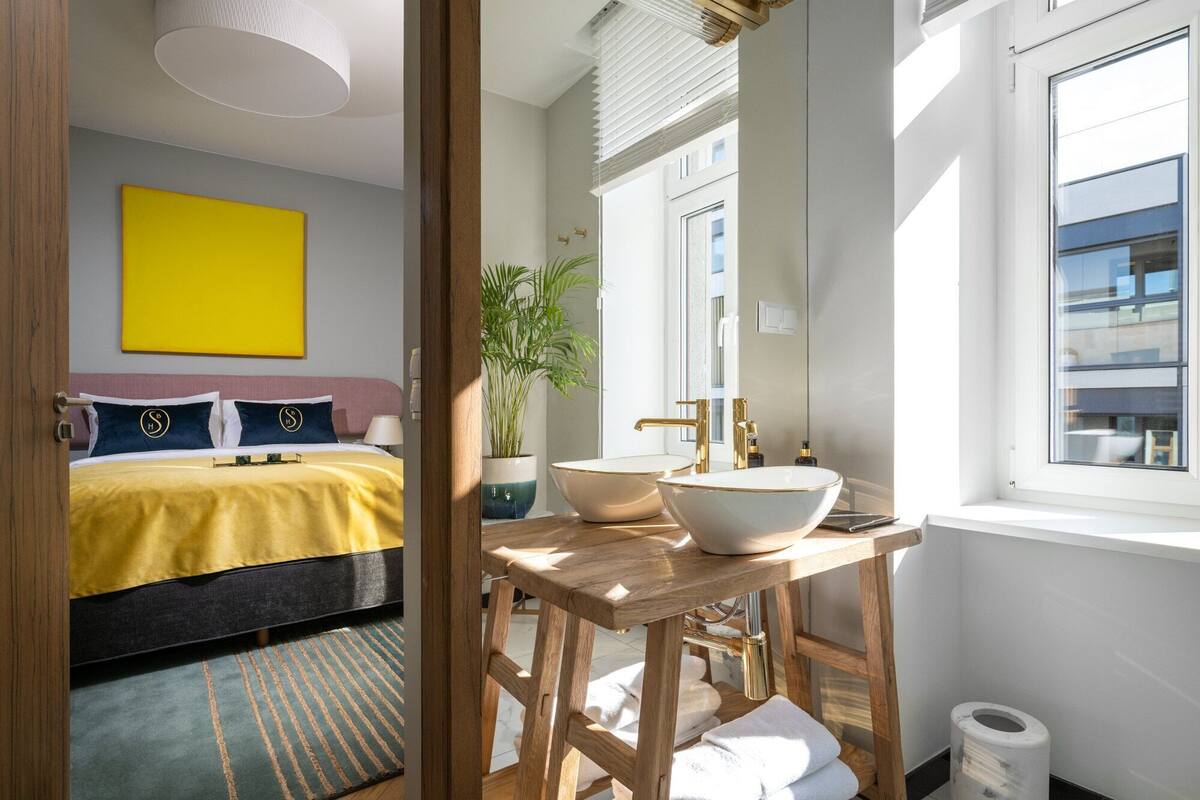 A modern hotel room with a bed, a large yellow artwork, a wooden side table with a bowl sink, and a view of the city through the window.