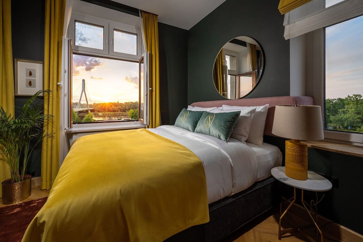 A bedroom with a bed, a round mirror, a bedside table with a lamp, and a window with curtains.