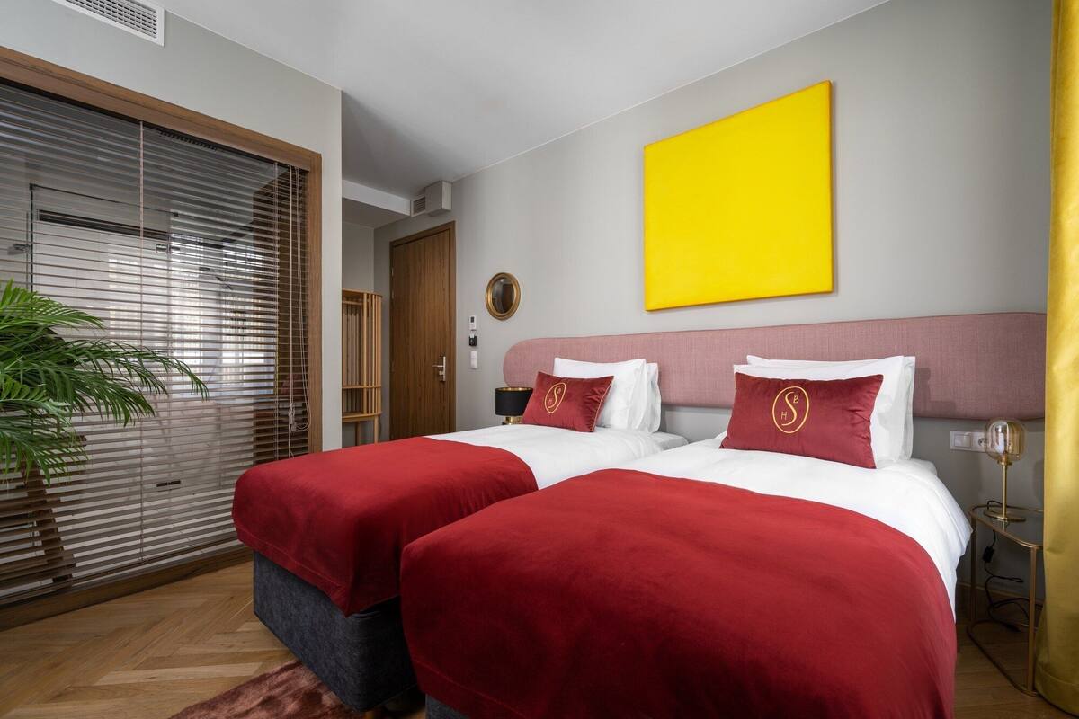 A hotel room with two beds, a large window with blinds, a yellow wall, and a plant.