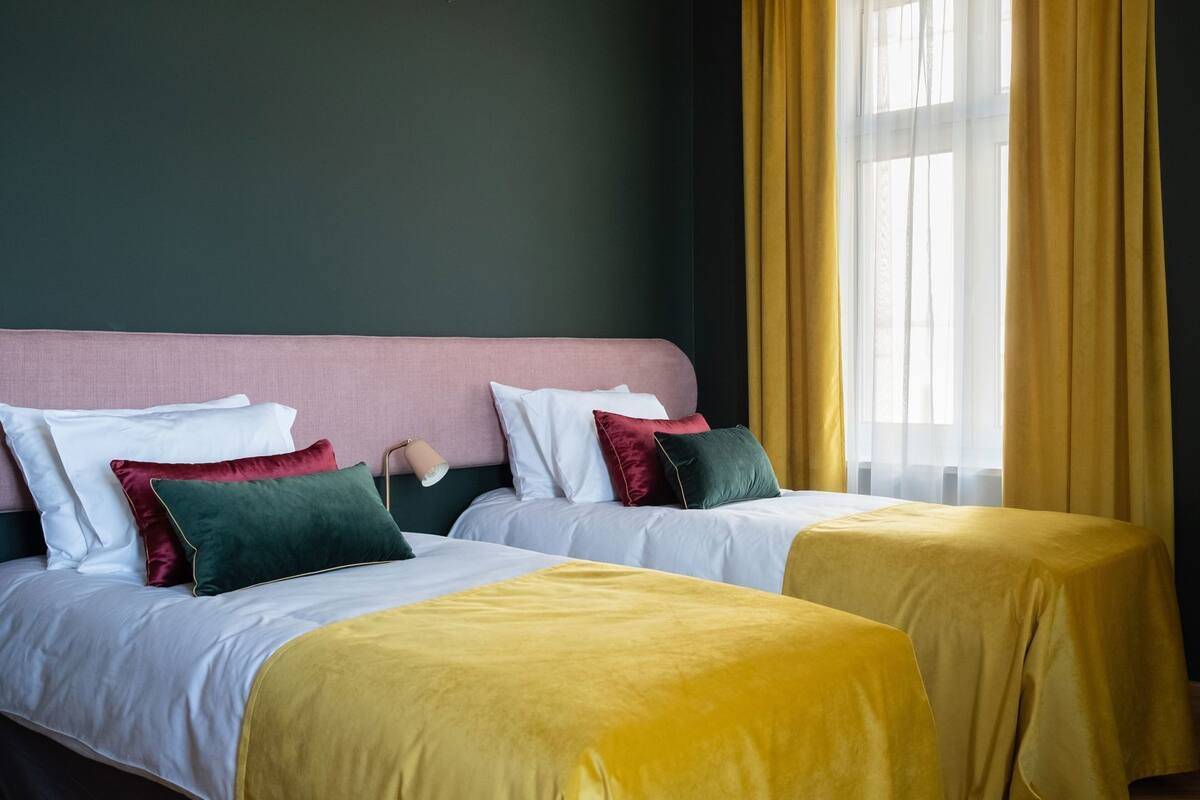 A hotel room with two beds, a nightstand, a lamp, and a window with curtains.