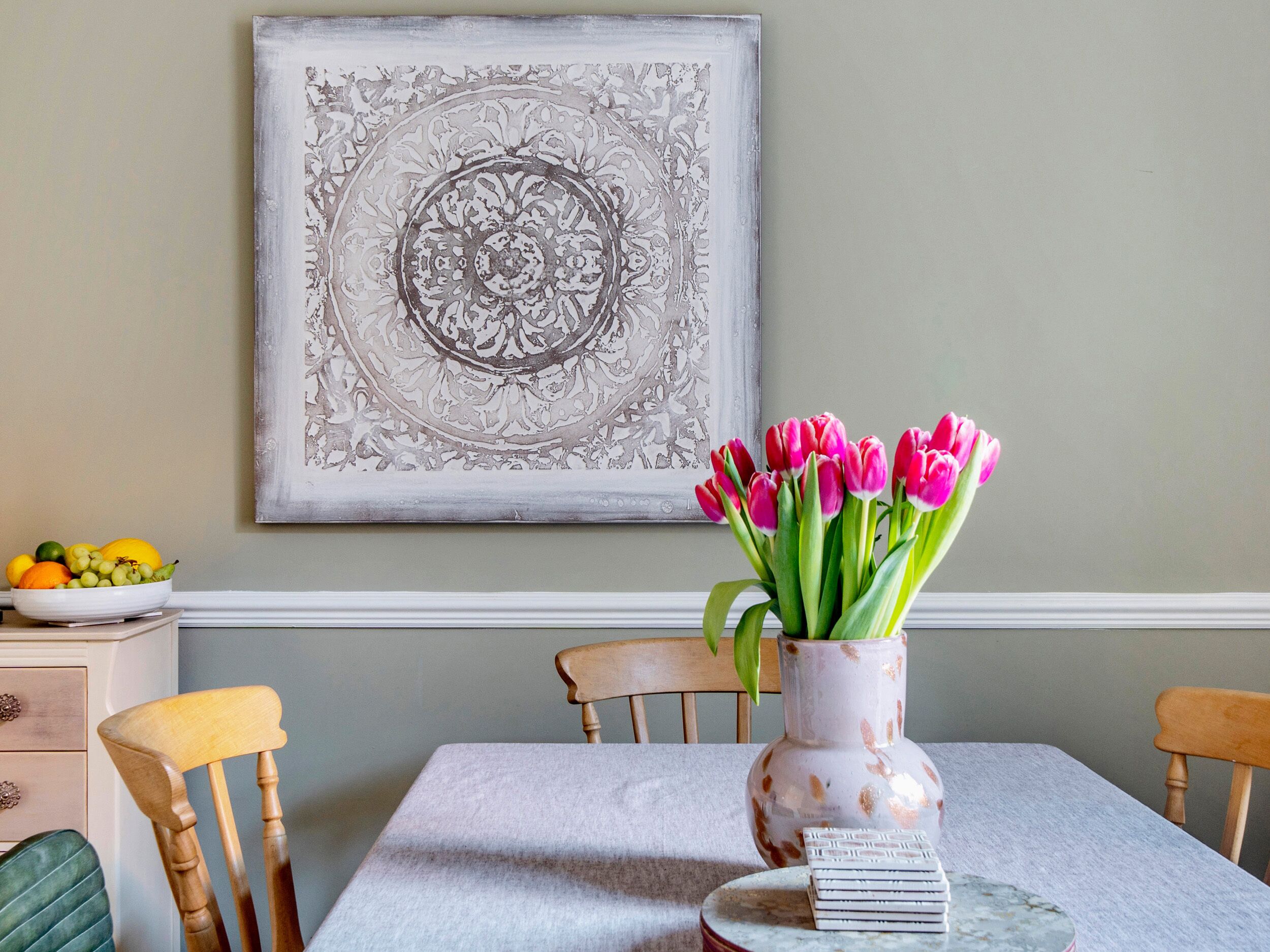 Et spisebord med en vase tulipaner, en skål med frugt og et indrammet kunstværk på væggen.