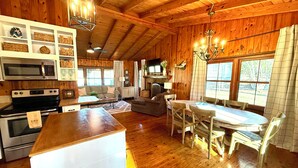 A cozy cabin interior with wooden walls, a kitchen area with a microwave and stove, a dining table with chairs, and a fireplace. - The Cozy Cabin on Lake Martin (Eclectic)