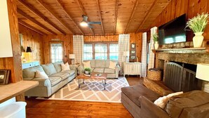 A cozy cabin-style living room with wooden floors, a fireplace, a television, and a large window. - The Cozy Cabin on Lake Martin (Eclectic)