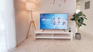 Una sala de estar con un televisor de pantalla plana sobre un soporte blanco, una lámpara de pie y una planta en maceta.