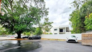 Un área de estacionamiento con un edificio blanco, un árbol y coches aparcados.