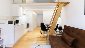 Una cocina moderna con armarios blancos, un microondas y una cafetera. Hay una zona de comedor con una mesa y sillas, y un sofá marrón en primer plano.