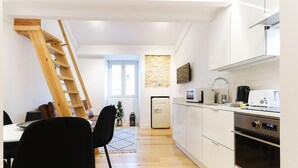 Una cocina moderna con armarios blancos, horno empotrado y microondas. Una escalera de madera está apoyada contra la pared, y se ve un comedor con una mesa y sillas al fondo.
