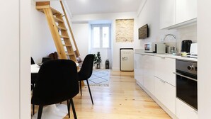 Cocina moderna con alacenas blancas, microondas integrado y horno de acero inoxidable. Se observa una escalera de madera apoyada en la pared y una zona de comedor con sillas negras y una mesa blanca.