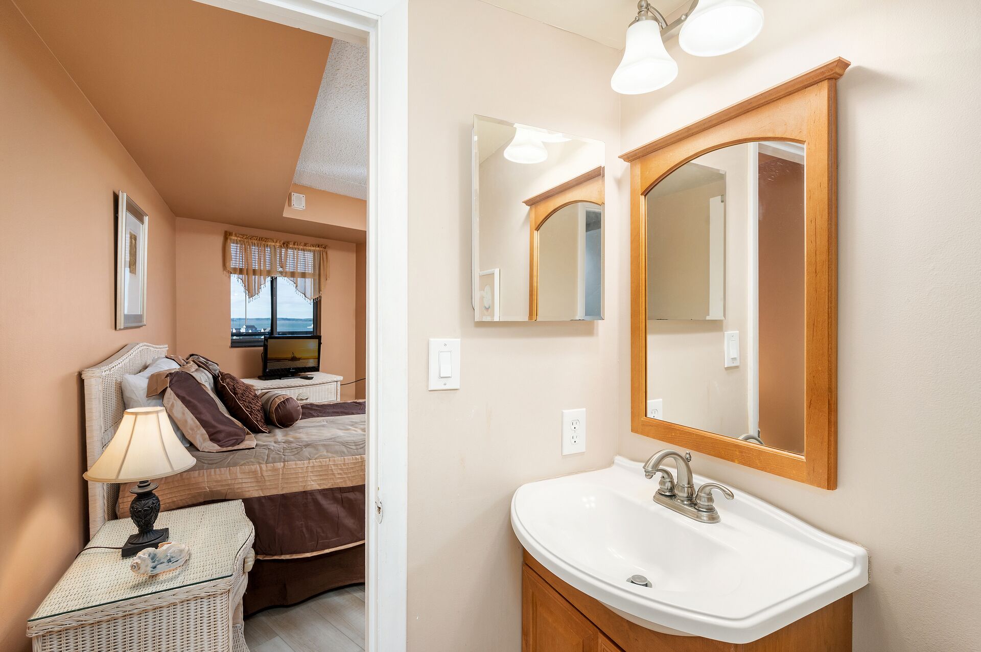 A bedroom with a bed, bedside lamp, and a television. There is a bathroom with a sink and mirror.