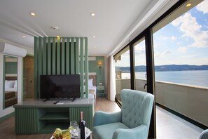 A balcony with a view of the sea, a TV on a stand, a light blue armchair, and a small table with a bottle and glasses. - ASPEN HOTEL ÇANAKKALE (Çanakkale)
