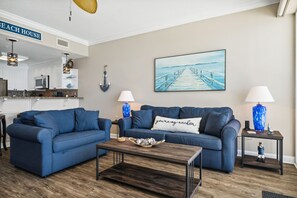 Een woonkamer met een blauwe zithoek, een salontafel en een schilderij van een pier.