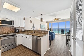 A modern kitchen with white cabinets, a granite countertop, stainless steel appliances, and a view of the ocean through large windows. - FREE Golf+Perks! Boardwalk Beachfront 1007 Beautifully Updated w/Heated Pool! (Panama City Beach)