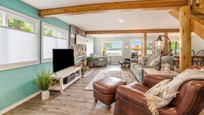 A spacious living room with a large TV, leather sofa, and wooden beams on the ceiling. - Family Fun on Otsego Lake - HUGE yard, kayaks, paddleboards, zip line and more! (Gaylord)