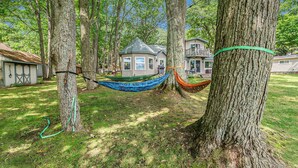 A hammock between trees in a wooded area with a house in the background. - Family Fun on Otsego Lake - HUGE yard, kayaks, paddleboards, zip line and more! (Gaylord)