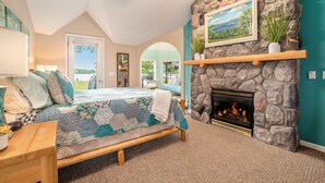 A bedroom with a stone fireplace, a bed with a quilt, a wooden bench, and a view through an arched doorway. - Family Fun on Otsego Lake - HUGE yard, kayaks, paddleboards, zip line and more! (Gaylord)
