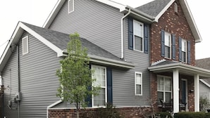 Une maison à deux étages, avec un revêtement extérieur gris, une partie en briques sur le côté droit et un petit porche à l’avant avec une chaise.