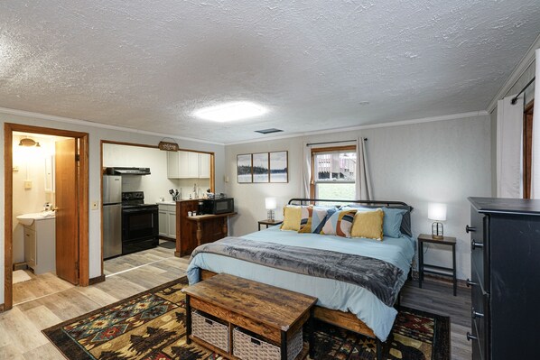 A bedroom with a bed, bedside tables, a dresser, a window with curtains, and a kitchenette in the background. - Riverside De Luxe*WiFi*Steps to the River (Elkins)