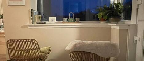 A room with a window sill, two wicker chairs, a white cushion, a potted plant, and a framed picture on the wall.