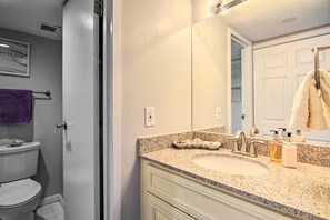 Un baño con inodoro blanco, lavamanos con encimera de granito y espejo sobre el lavamanos.