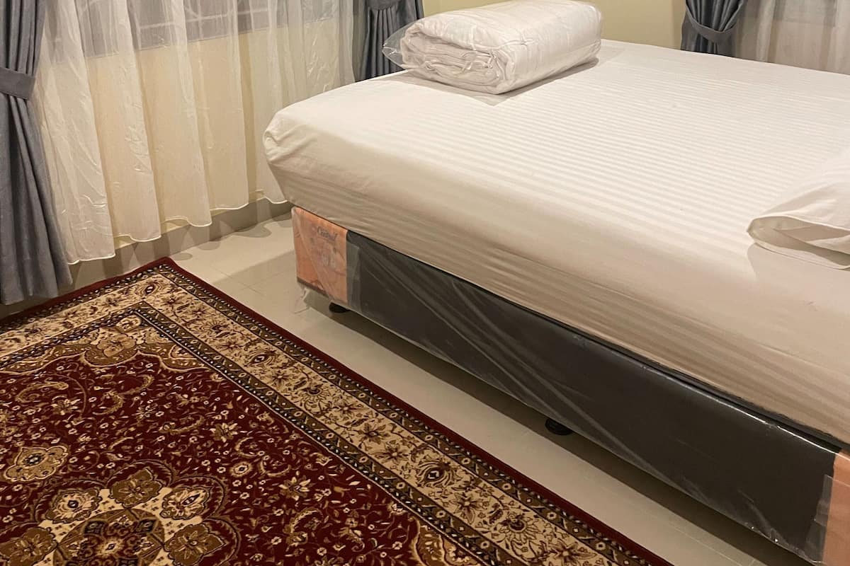 Una cama bien tendida con sábana blanca y toalla doblada, un tapete estampado y una ventana con cortinas de gasa.