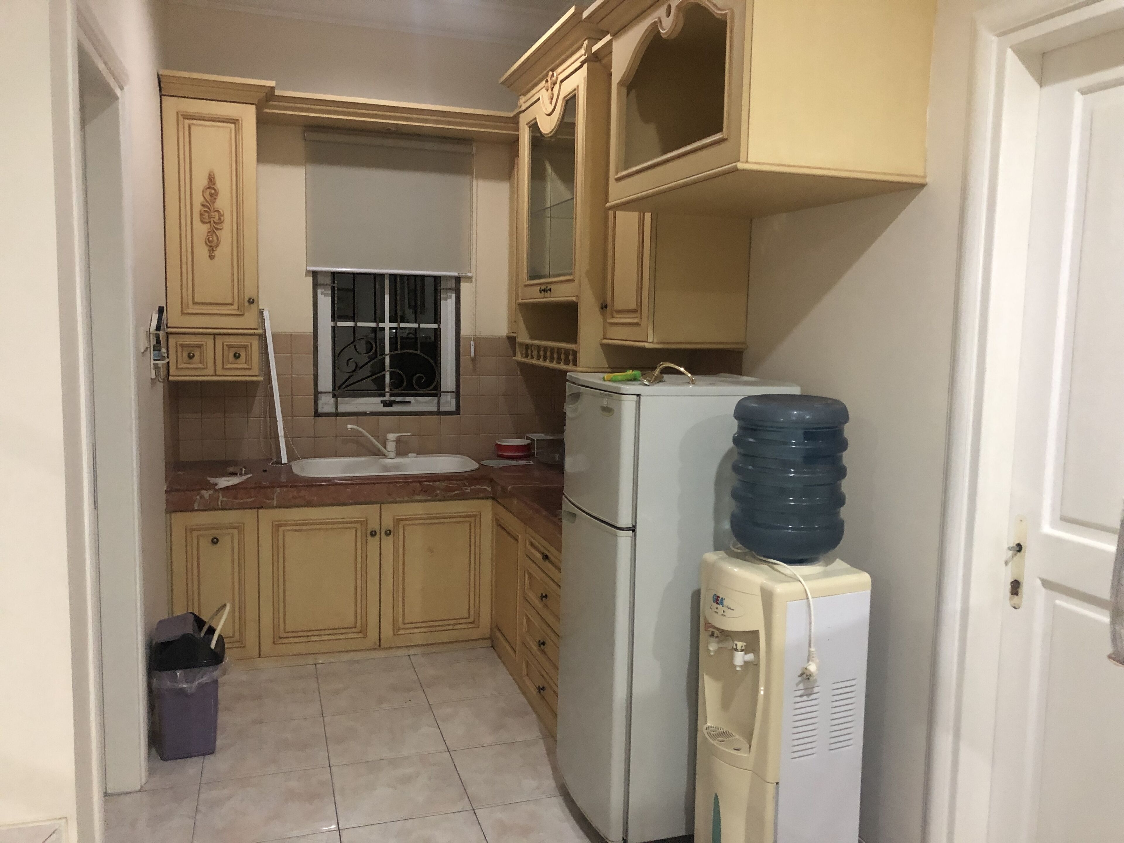 Una cucina compatta con mobili beige, un lavello, un frigorifero e un dispenser di acqua.