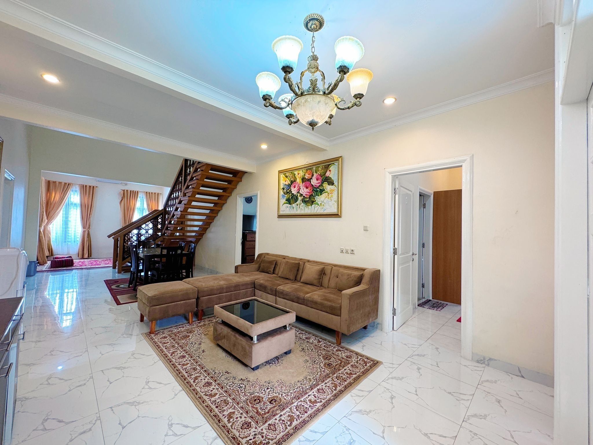 A spacious living area with a large sectional sofa, a glass coffee table, and a patterned rug. A staircase leads to another level, and a chandelier hangs from the ceiling.