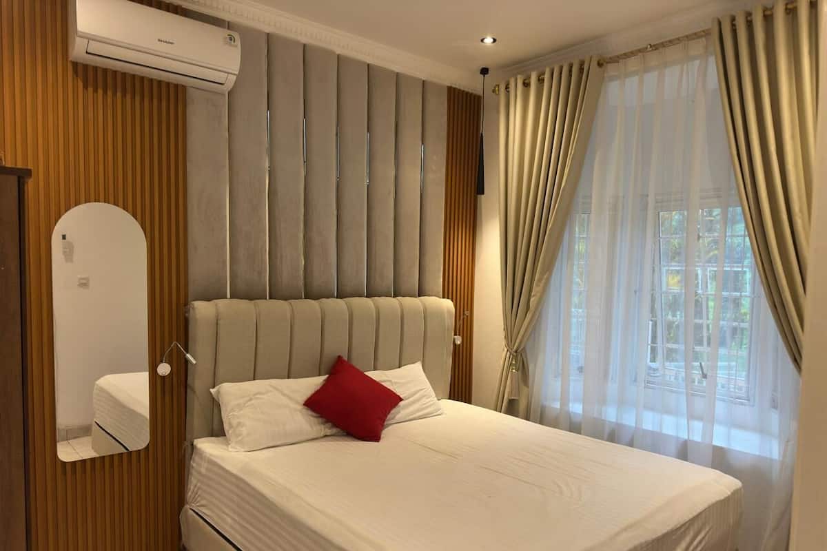 Un dormitorio moderno con una cama grande, cabecera de listones verticales, un espejo junto a la cama y una ventana con cortinas traslúcidas.