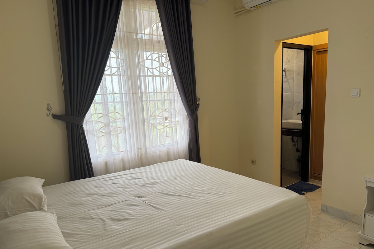 Hotelkamer met een bed, gordijnen, een raam met uitzicht, een zichtbare badkamer via een geopende deur, en een betegelde vloer.