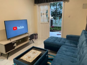 Een woonkamer met een flatscreen-tv waarop het YouTube-logo te zien is, een blauwe bank, een glazen salontafel met een decoratief dienblad, en een deur die leidt naar een balkon met planten.
