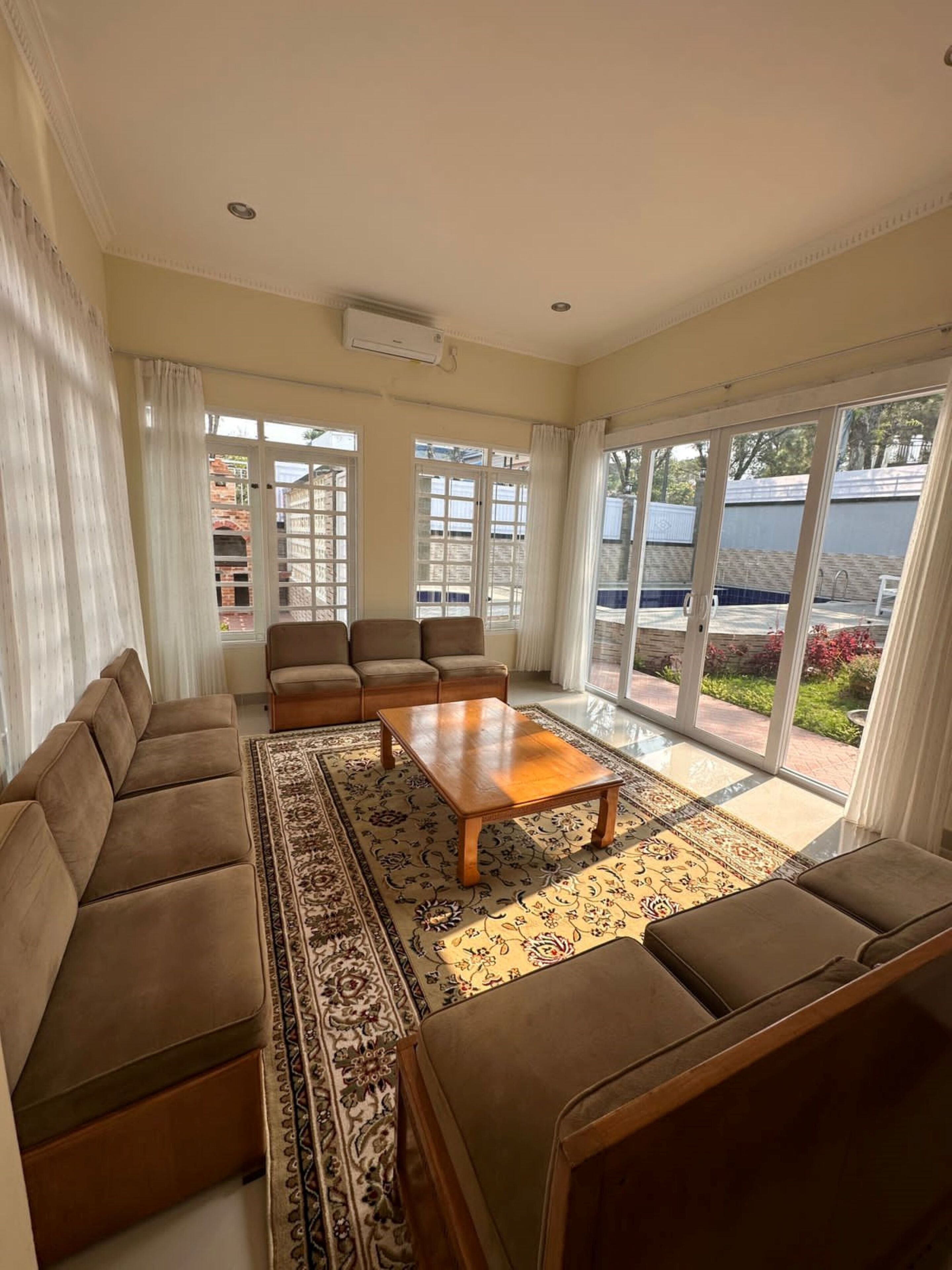A spacious living room with a sofa set, a coffee table, and a large window with a view of a swimming pool and greenery.