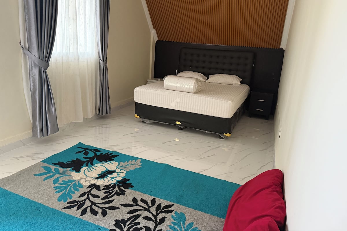 Un dormitorio con cama, cortinas, cabecero y alfombra estampada.