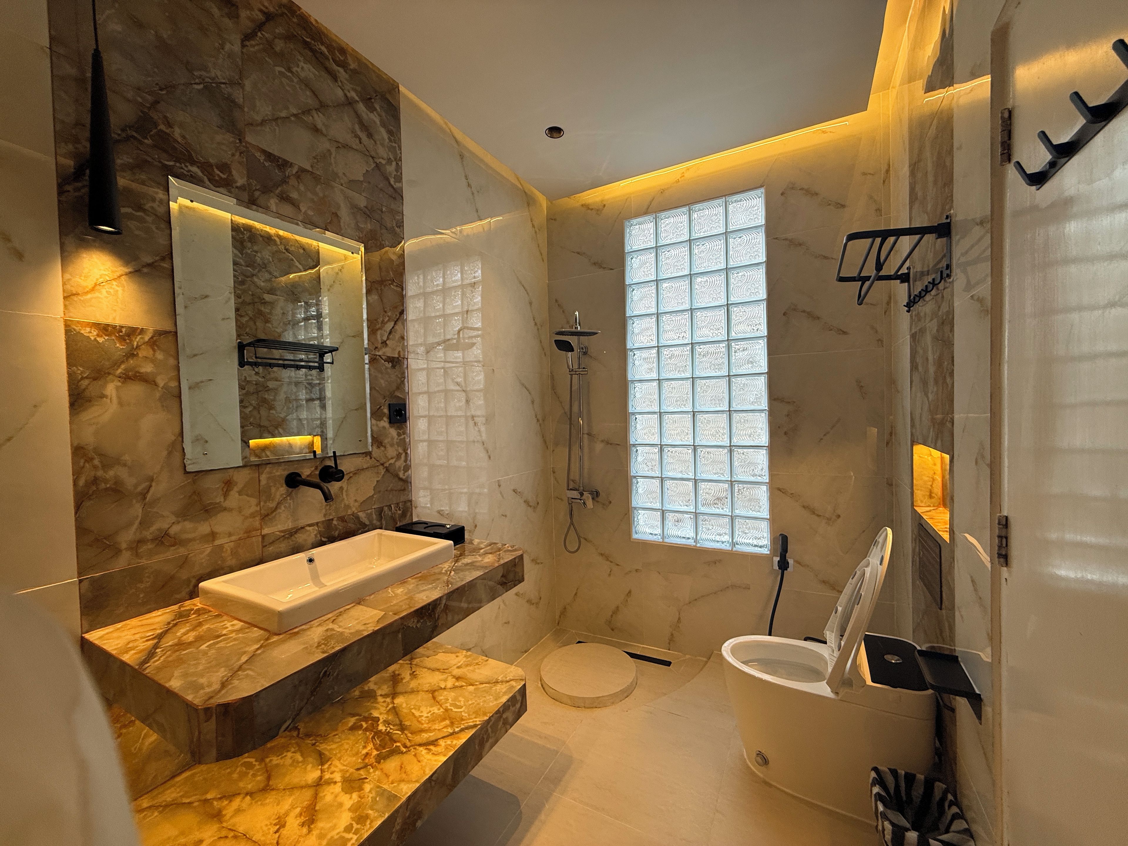 A modern bathroom with a marble vanity, a large mirror, a shower, and a window with decorative glass panels.
