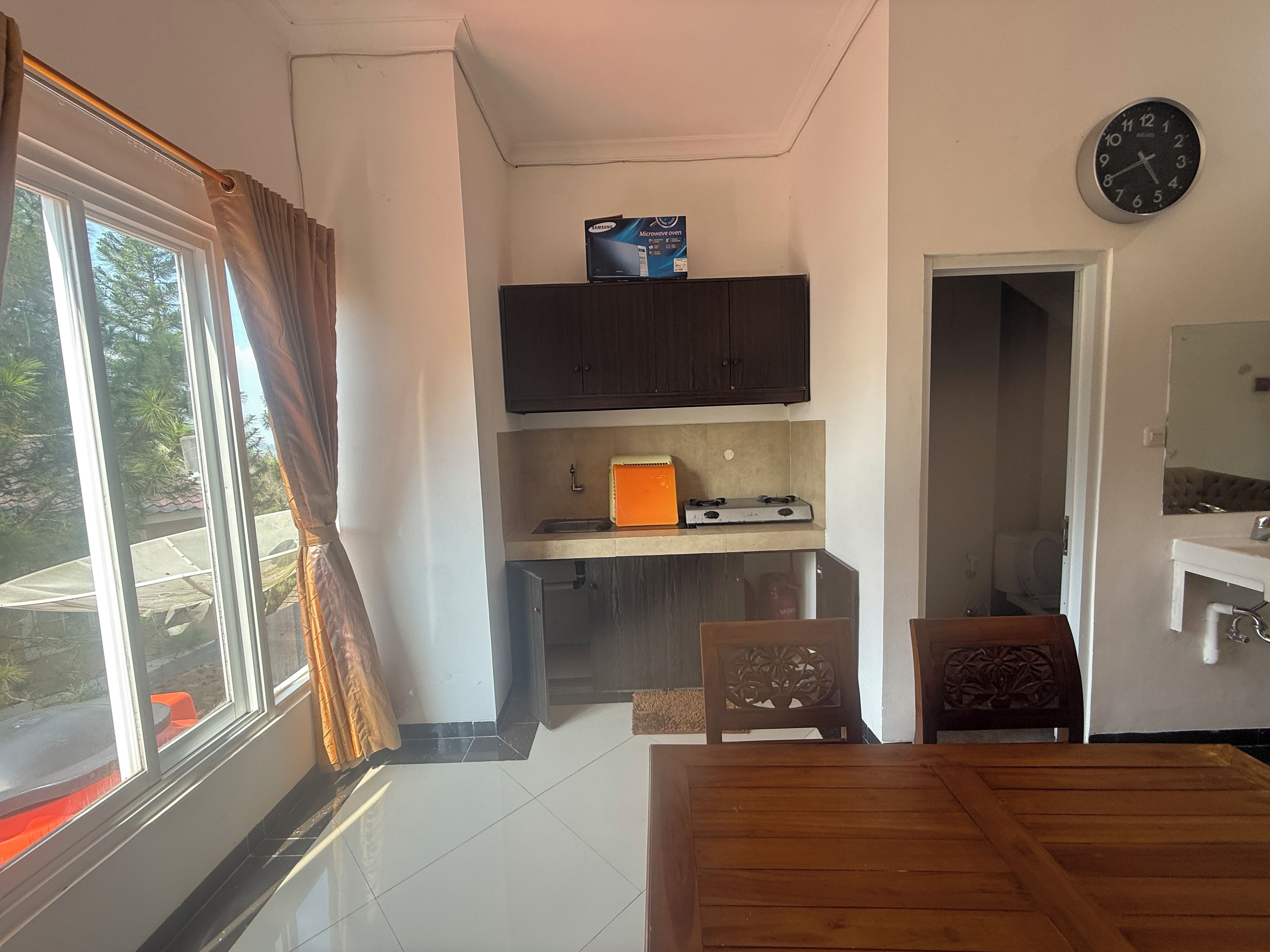 Una cocina compacta con microondas, un pequeño electrodoméstico naranja y un armario. Hay una mesa de comedor con sillas, un reloj en la pared y una ventana con cortinas.