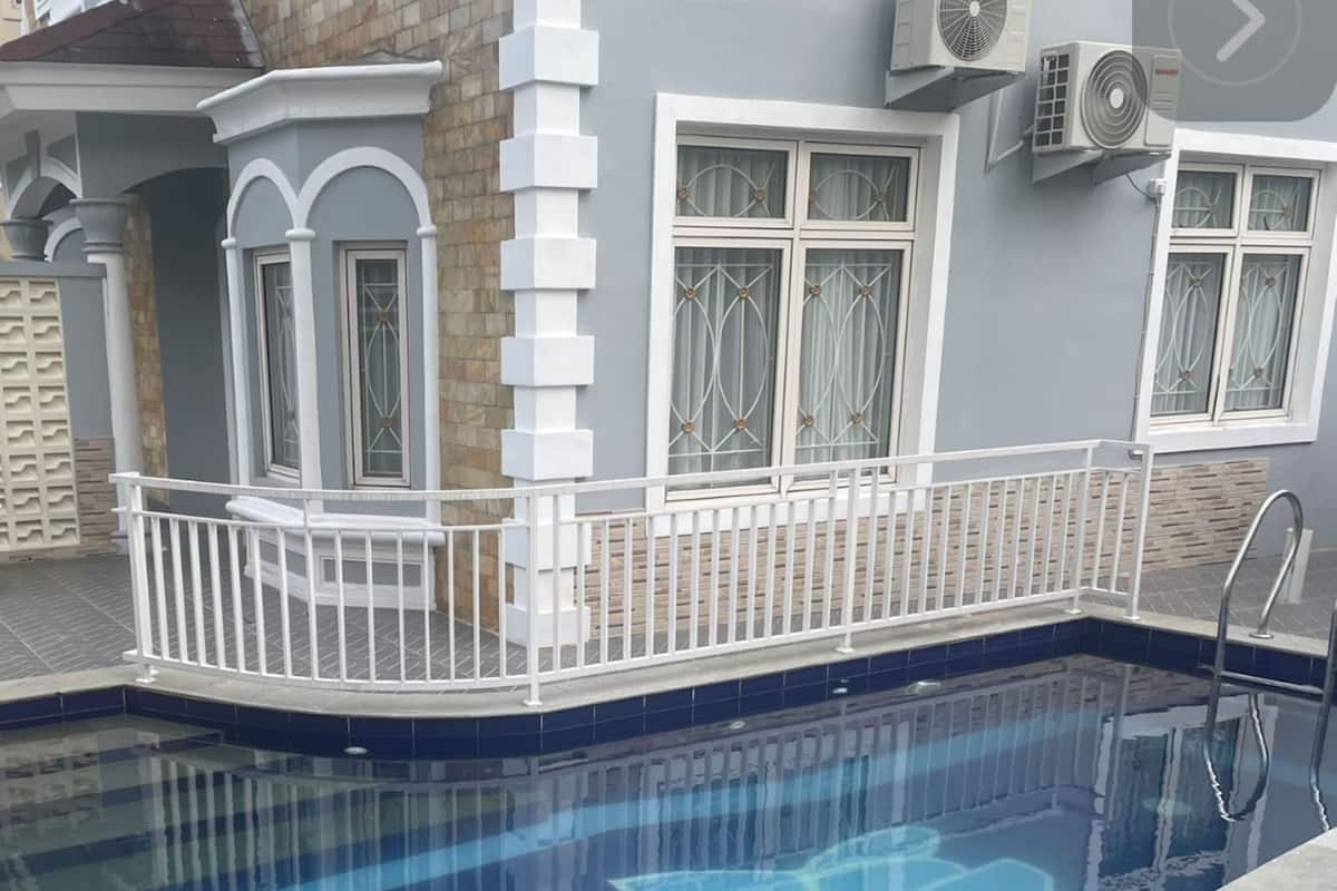 Una piscina con diseño geométrico, rodeada por una valla blanca y una casa con fachada de piedra.