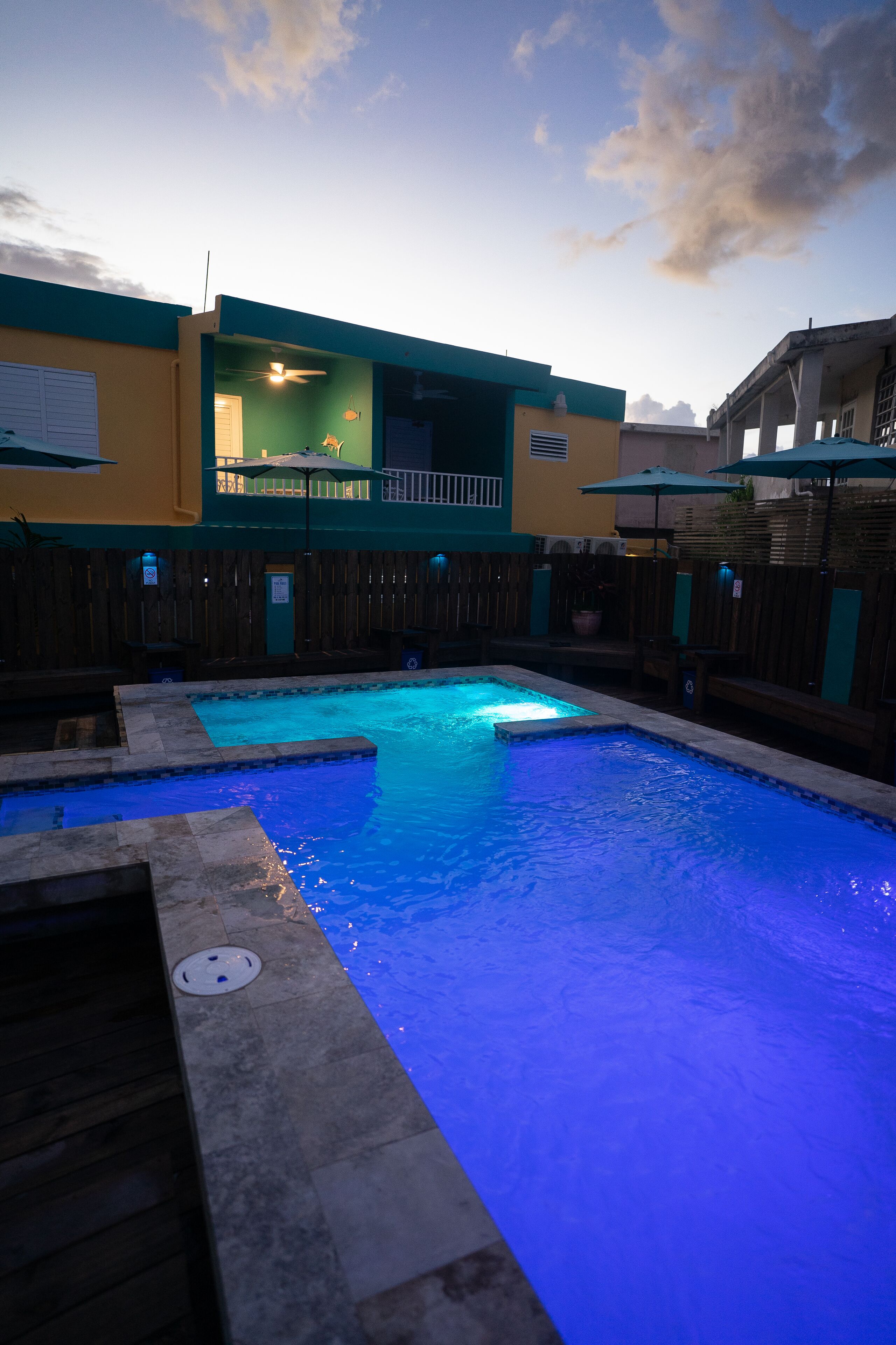 A swimming pool with a blue-lit edge, surrounded by a wooden fence and a building with a balcony.