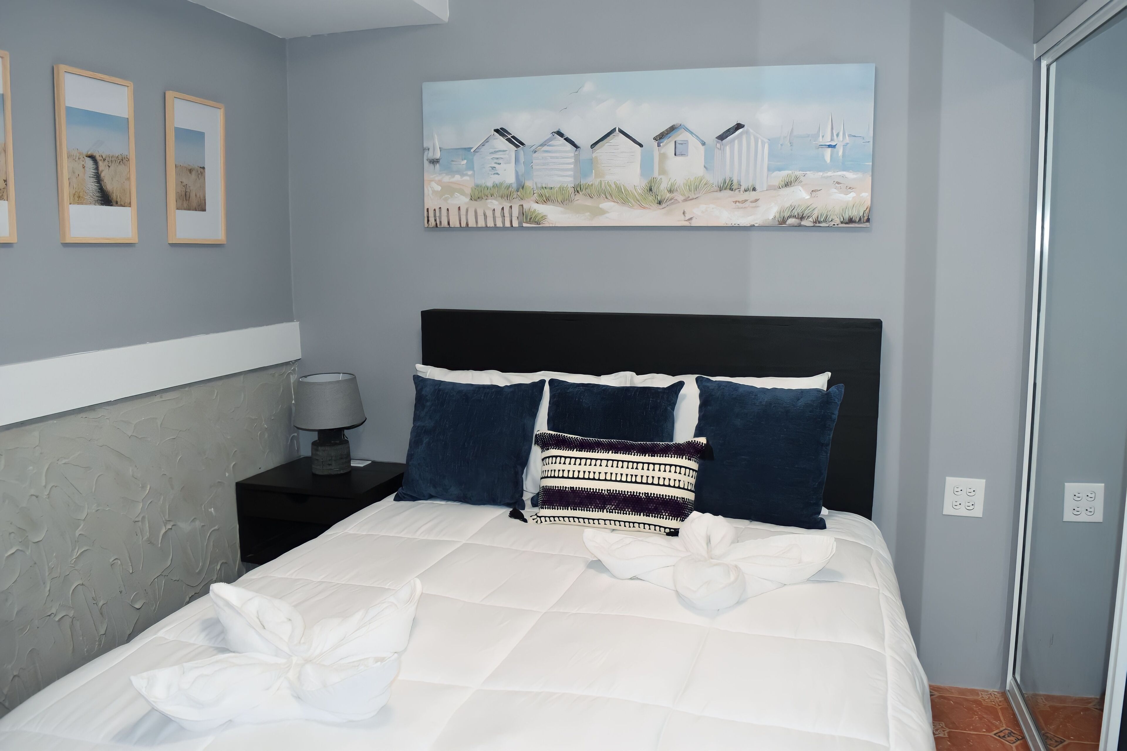 A neatly made bed with white linens and a decorative pillow, a bedside table with a lamp, and a painting of beach huts on the wall.