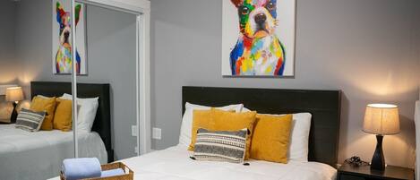 Ein Hotelzimmer mit einem großen Bett, einem Nachttisch mit Lampe, einem kleinen Tisch mit Korb und zwei Wandbildern von Hunden.